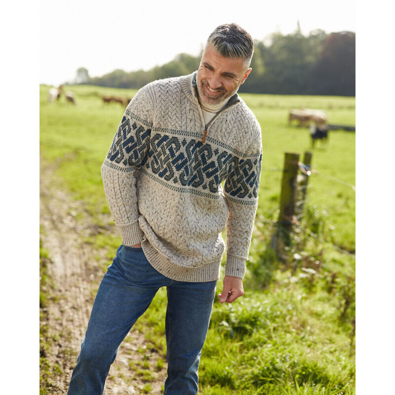 Man standing in a grass field wearing an oatmeal coloured Aran cable knit half zip jacquard sweater